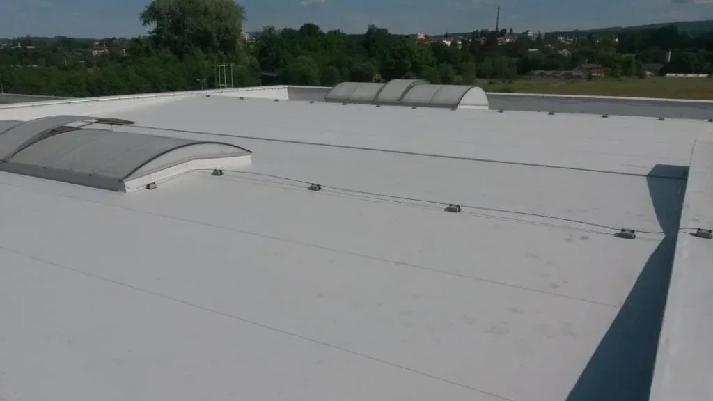 Střecha haly s obloukovým světlíkem, za ní zelené pole a město, slunečno