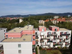 Zvýšený pohled na bytový dům s korálovou fasádou, dvě střešní plochy přitížené kačírkem, v pozadí Ještěd