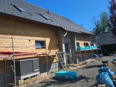 Sedlová střecha s tmavou skládanou krytinou a třemi střešními okny nad fasádou s dřevěným obkladem, podél domu lešení