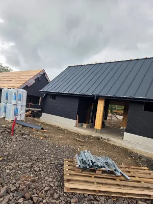 Novostavba s tmavou fasádní fólií a sedlovou střechou z tmavého plechu na stojatou drážku, před domem palety a stavební materiál