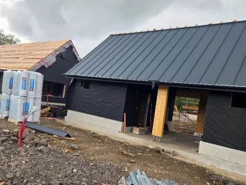 Novostavba s tmavou fasádní fólií a sedlovou střechou z tmavého plechu na stojatou drážku, před domem palety a stavební materiál
