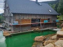 Dlouhá strana sedlové střechy s tmavou krytinou, komínem s lávkou a střešními okny, pod ní fasáda v lešení a jezírko