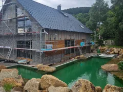 Dlouhý dům se sedlovou střechou s tmavou krytinou, prosklený štít a před domem zahradní jezírko s balvany